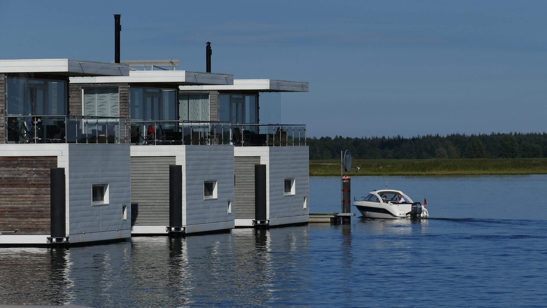 A row of houseboats fixed to dock pilings, serving as a floating pier, with a small yacht approaching for landing.